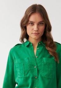 Cămașă verde cu nasturi, fabricată dintr-un material neted, având două buzunare pe piept și guler. Mâncile sunt ușor suflecate.