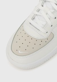 Λευκό δερμάτινο αθλητικό παπούτσι με διάτρητο γκρι μέρος στη μύτη, λευκά κορδόνια και ασημί λογότυπο Nike. Διαθέτει ομαλή υφή και δομημένη σόλα.