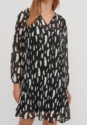 Sort plisseret kjole med hvidt abstrakt penselstrøgsmønster, V-hals og lange transparente ærmer, båret af en stående person.