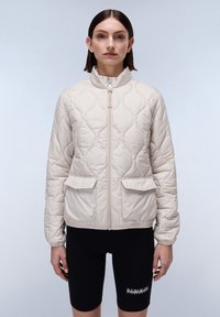 Beige gewatteerde jas met een hoge kraag en een rits aan de voorkant, met twee vo pockets en elastische manchetten, gedragen over zwarte shorts.