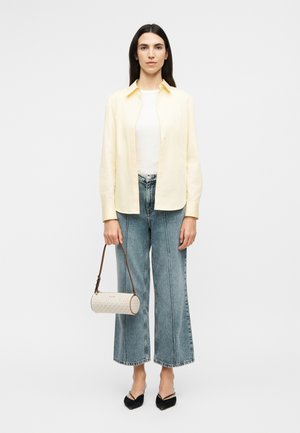 Woman with long dark hair wearing a pale yellow shirt, white top, wide-leg blue jeans, black shoes, holding a small round white handbag.