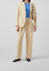 Beige maßgeschneiderter Anzug mit lockeren Hose, einem blauen Rippoberteil und schwarzen Slippern. Schlichte Handtasche mit einem Riemen.