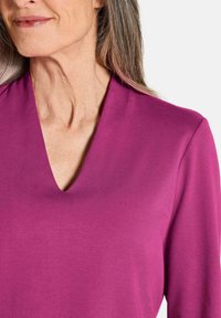 Top a manica lunga con scollatura a V in magenta brillante. Tessuto liscio e morbido con una leggera lucentezza. Design semplice, senza motivi o accessori visibili.