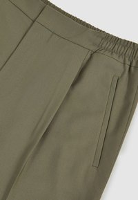 Pantalon vert olive en tissu texturé. Dispose d'une taille élastique et de poches latérales, avec des lignes épurées et des détails de couture minimaux.