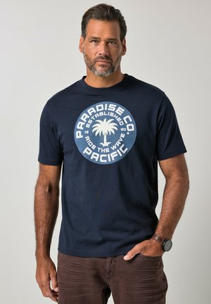 Uomo con barba che indossa una maglietta blu navy con logo di palma bianco e testo "Paradise Co Pacific" sta in piedi davanti a uno sfondo semplice.