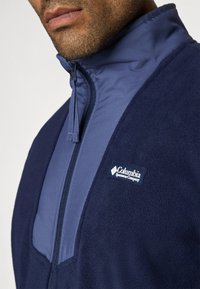 Tumesinine fliisjakker kõrge kraega, lukuga ja helesiniste aktsentidega. Rinnal on Columbia logo. Pehme tekstuur.