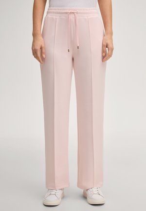 Femme portant un pantalon large rose clair avec des coutures frontales et une taille à cordon, associé à des baskets blanches et un haut blanc.