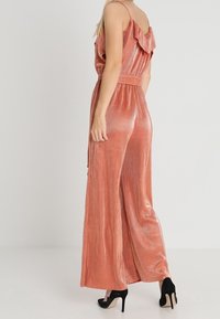 Jumpsuit de oro rosa con detalle de hombros con volantes, piernas fluidas y cintura con cinturón, combinado con tacones altos negros. Textura suave y brillante.