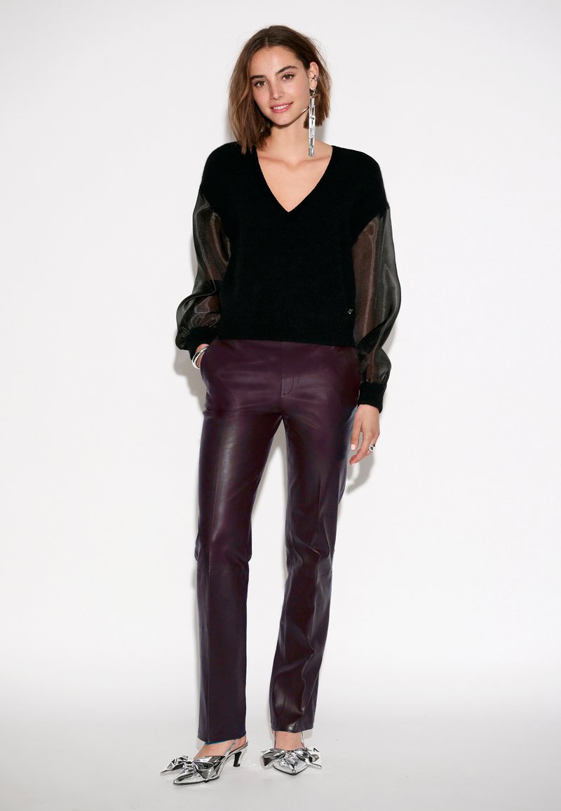 Zwarte gebreide trui met doorschijnende pofmouwen, gecombineerd met strakke donkerpaarse leren broeken en metallic zilveren hakken met strikaccenten.