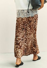 Personne portant une jupe maxi marron et blanche à pois avec une bordure en dentelle, des chaussures noires sans lacets, et tenant un sac en cuir noir.