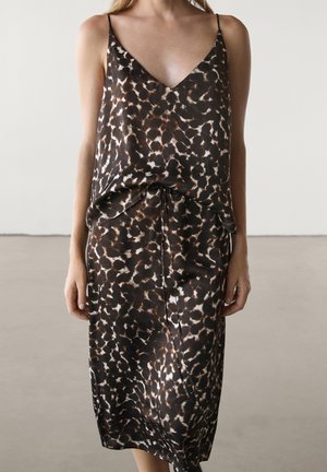Woman wearing sleeveless leopard print slip dress with thin straps and a tie waist, standing against a plain background.