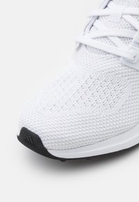 Hvide sportsko med en mesh-overdel, tekstureret overflade og en sort gummisål. Har hvide snørebånd og et Adidas-logo på siden.