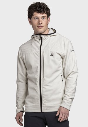 Junger Mann trägt eine hellbeige Kapuzenjacke mit schwarzem Reißverschluss und Logos, steht vor einem schlichten hellen Hintergrund.