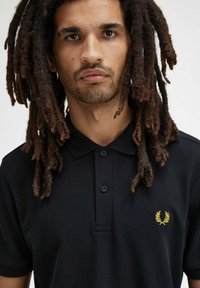 Polo nero con tessuto a trama, dotato di una patta con due bottoni e un piccolo logo a alloro giallo sul petto sinistro.