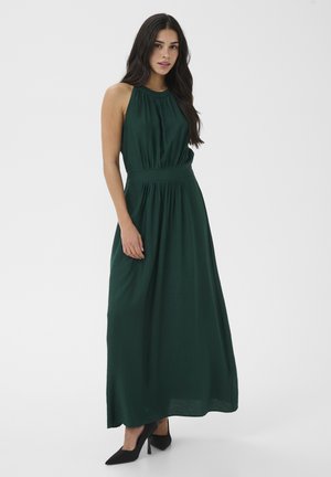 Donna che indossa un abito lungo verde scuro senza maniche con collo all'americana e vita arricciata, in piedi con una mano rilassata lungo il fianco e scarpe con tacco nere.