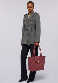 Szara wełniana kurtka z czterema kieszeniami i paskiem, połączona z czarnymi spodniami. Trzyma bordową skórzaną torbę na ramię z mosiężną klamrą.