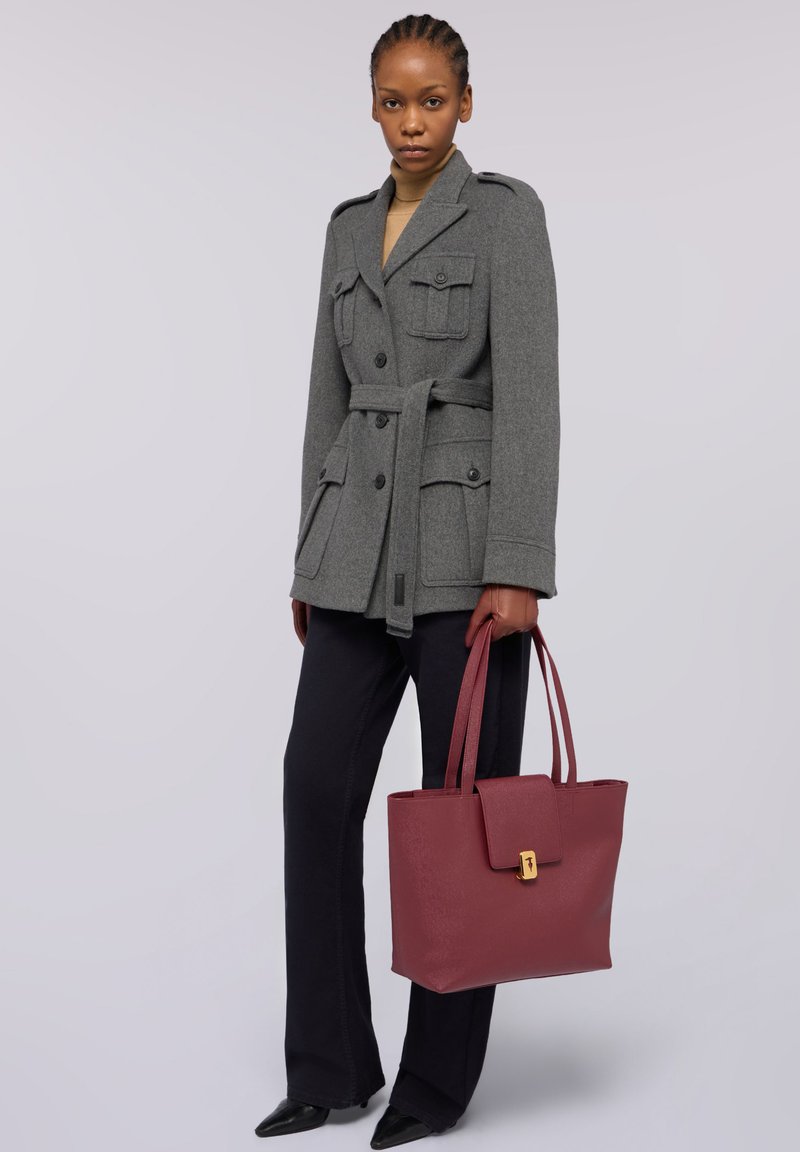 Szara wełniana kurtka z czterema kieszeniami i paskiem, połączona z czarnymi spodniami. Trzyma bordową skórzaną torbę na ramię z mosiężną klamrą.