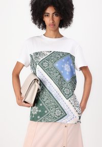T-shirt blanc avec des patches aux motifs variés en vert, bleu et blanc ; présente des designs de bandana et de zèbre. Le modèle tient un sac à main beige.