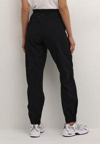 Czarne dresowe spodnie na elastycznej talii z zwężanymi mankietami. Tkanina wygląda na lekką i fakturowaną, w połączeniu z białymi i szarymi sportowymi butami.