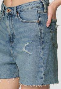 Jasnoniebieskie szorty dżinsowe z postrzępionymi brzegami, z przetartym obszarem na kolanie i kieszeniami z przodu. Mosiężne akcenty sprzętowe dopełniają design.