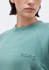 Κοντινό πλάνο ενός ατόμου που φοράει ένα τιρκουάζ φούτερ με το κέντημα "BORG" στο στήθος, εμφανίζοντας τον λαιμό και το κάτω μέρος του προσώπου.