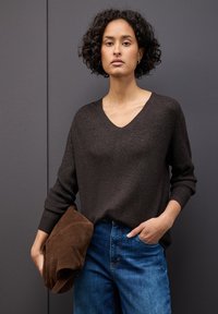 Kobieta z krótkimi, kręconymi włosami, ubrana w ciemnobrązowy sweter z dekoltem w serek i niebieskie dżinsy, trzymająca brązowy zamszowy kapelusz, na tle szarej ściany.