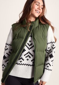Olive green puffer vest with a stand collar, unzipped over a light gray sweater featuring black geometric patterns and ribbed cuffs.