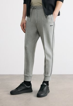 Mees, kes kannab halli BOSS dressipükst mustade küljeribadega, musti tossusid ja musta jopet, üks käsi taskus.