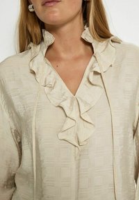 Light beige blouse featuring a textured fabric, v-neckline, and ruffled collar with ties. Soft, flowing material with a slight sheen.