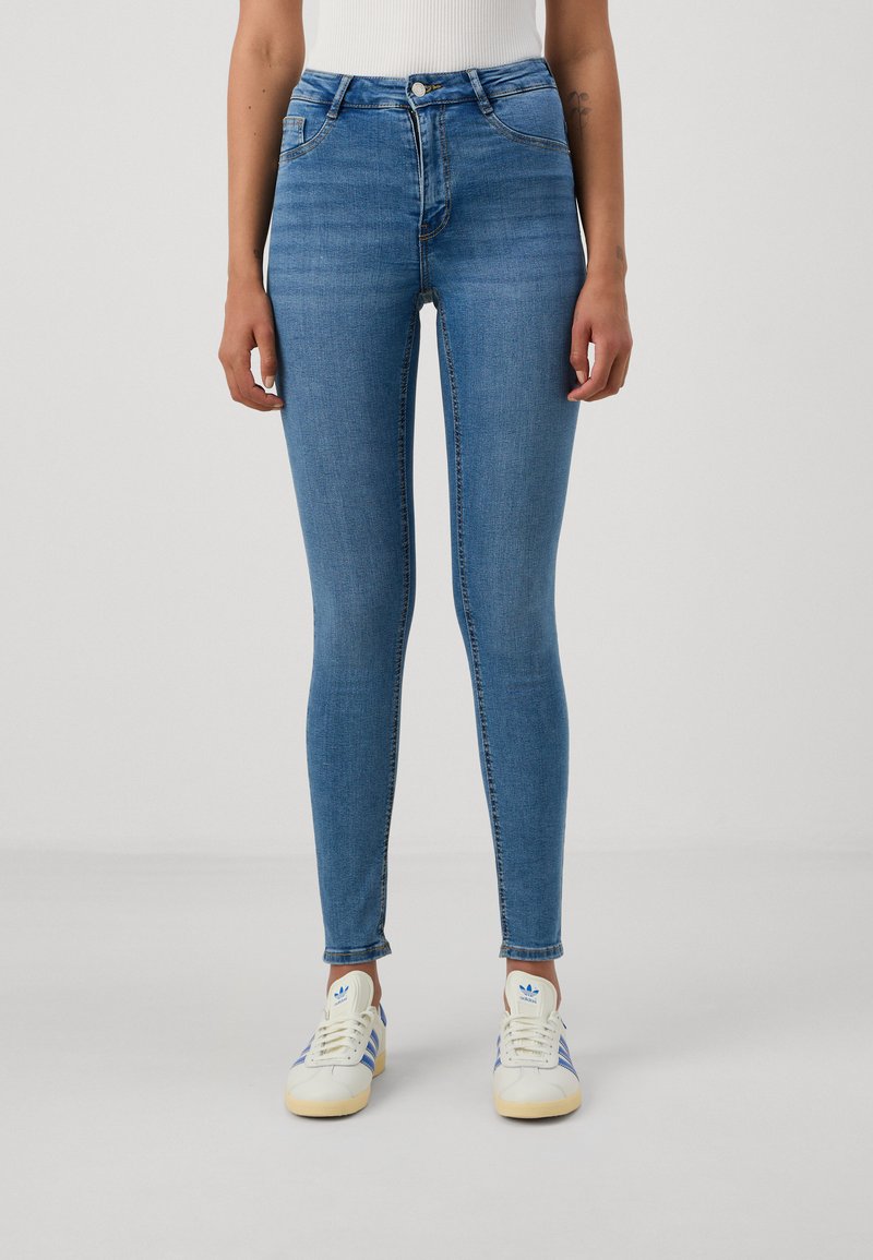 Skinny blue denim jeans with a high waist, featuring a zip and button closure, two front pockets, and narrow leg openings, paired with white sneakers.