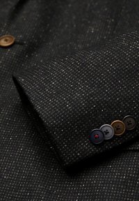 Dunkles gemustertes Gewebe eines Jackets, das eine strukturierte Oberfläche mit wechselnden Farbtönen aufweist. Vier Knöpfe in Braun, Schwarz, Grau und einem roten Akzent.