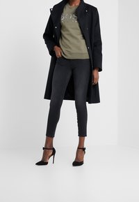 Donna che indossa un lungo cappotto nero, una felpa verde oliva di Hugo Boss, jeans skinny neri e scarpe con tacco alto e cinturino alla caviglia nere.