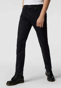 Jeans de mezclilla negra con un corte ajustado, con un diseño de cinco bolsillos, costuras sutiles y un pequeño cierre de botón metálico. Combinados con botas negras.