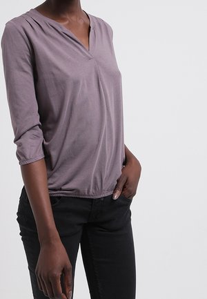 Frau trägt ein mauvefarbenes Langarmshirt mit V-Ausschnitt und schwarze Hose, eine Hand in der Tasche, vor einfachem hellem Hintergrund.