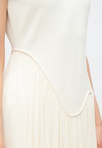 Kremowa sukienka z gładką górną częścią i teksturowaną, lejącą się tkaniną poniżej, z asymetrycznym dołem i marszczonymi akcentami.