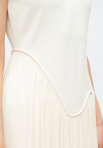 Κρεμ φόρεμα με λείο άνω τμήμα και υφασμάτινη υφή στο κάτω μέρος που ρέει, με ασύμμετρο στρίφωμα και συγκεντρωμένες λεπτομέρειες.
