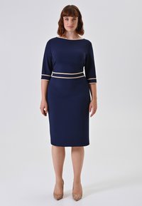 Frau, die in einem knielangen, marineblauen Kleid mit Dreiviertelärmeln und beigem Saum an der Taille und den Ärmelmanschetten steht, und beigen hohen Schuhen.