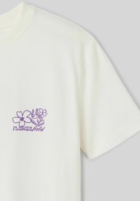 T-shirt en coton couleur crème avec un graphisme floral violet et le texte "CHAMP DE FLEURS" sur la poitrine gauche. Manches courtes, coupe décontractée.