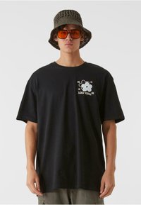 Črna bombažna majica s sproščenim krojem, ki ima grafičen cvetlični dizajn in napis na prsih. Oblečena z teksturirano vedro kapo in oranžnimi sončnimi očali.