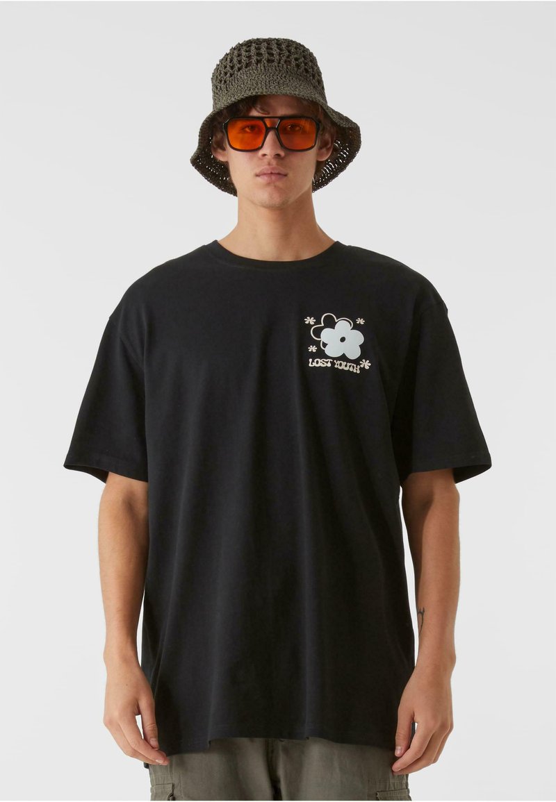 Črna bombažna majica s sproščenim krojem, ki ima grafičen cvetlični dizajn in napis na prsih. Oblečena z teksturirano vedro kapo in oranžnimi sončnimi očali.