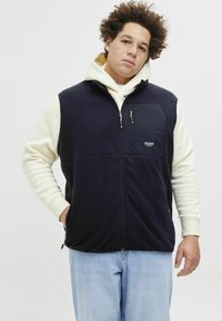 Granatowy fleece'owy kamizelka z zamkiem z przodu, z kieszenią na piersi i kieszeniami bocznymi, noszona na białym hoodie, w zestawieniu z jasnymi dżinsami.