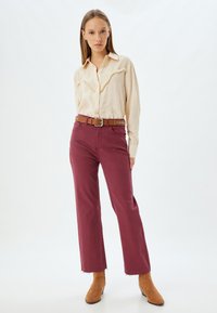 Camicia beige a maniche lunghe con dettagli in cucitura testurizzati, abbinata a pantaloni a gamba larga color bordeaux e una cintura beige con borchie argentate. Stivaletti marroni.