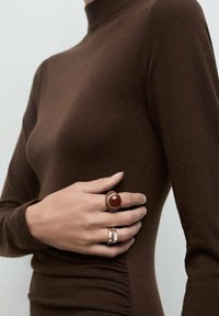 Maglione aderente a collo alto marrone abbinato a due anelli: uno con una pietra rossa e dettagli in oro, l'altro con un accento rettangolare in argento.