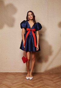 Φόρεμα από σατέν μπλε ναυτικό με φουσκωτά μανίκια και κόκκινο φιόγκο στη μέση, συνδυασμένο με κόκκινη τσάντα με χάντρες σε σχήμα καρδιάς και λευκές μυτερές μπαλαρίνες.
