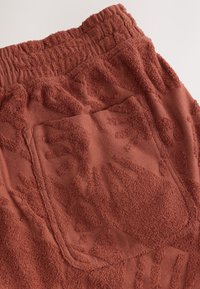 Gros plan d'un tissu texturé couleur rouille avec un motif en relief de soleil et de formes abstraites et une ceinture élastique.