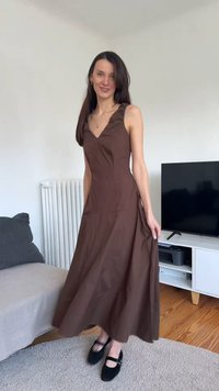 Γυναίκα με μακριά σκούρα μαλλιά, ντυμένη με καφέ αμάνικο μακρύ φόρεμα και μαύρες παπούτσια Mary Jane, στέκεται σε ένα σαλόνι.