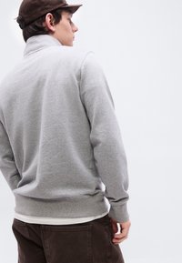 Νεαρός άνδρας φοράει γκρι φούτερ, λευκό εσώρουχο, σκούρο καφέ παντελόνι και σκούρο καφέ κάπα, κοιτώντας πλαγίως σε απλό φόντο.