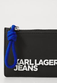 Juodas užtrauktuku užsidarantis maišelis su baltais žodžiais "KARL LAGERFELD JEANS". turi mėlyną tekstūruotą virvę, kad būtų lengviau laikyti. Sklandus gumos paviršius.