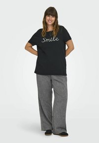 Juoda medvilninė marškinė su tekstu "Smile", pagamintu iš akmenukų, derinami su šviesiai pilkomis plačiomis kelnėmis iš tekstūruoto audinio ir juodais lengvais bateliais.