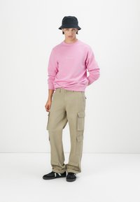Sweatshirt rose avec col rond et détail brodé, associé à un pantalon cargo beige avec plusieurs poches et des baskets noires. Chapeau seau noir.